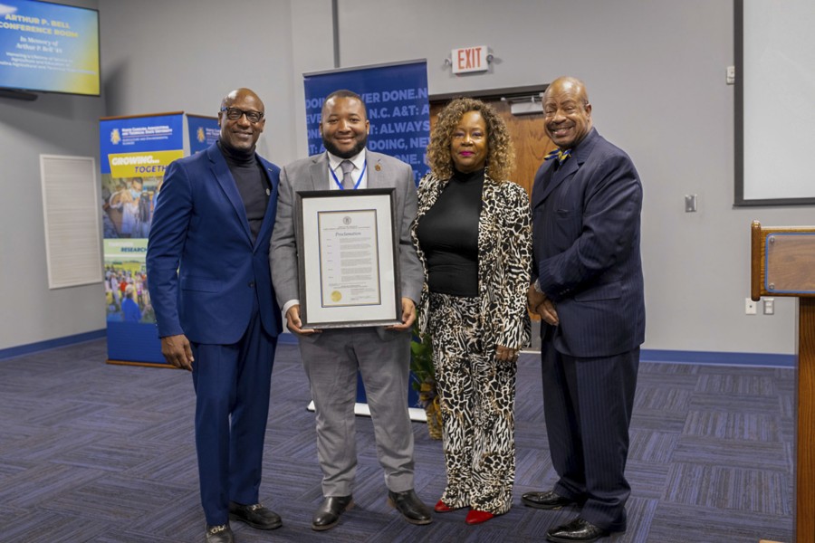 Four professionally dressed individuals pose in a conference room, smiling at the camera. The man in the center holds a framed proclamation document. To his left, a man in a navy blue suit and black turtleneck stands with his hand at his side. To his right, a woman in a black top and a black-and-white patterned pantsuit stands with her hands at her sides. On the far right, a man in a dark pinstripe suit and bow tie stands with his hands clasped. The background features banners with text related to North Carolina A&T State University and a wooden podium.