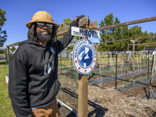 Smiling man with dreadlocks and sunglasses stands next to a sign that reads "The McKoy Way" and features the "Homegrown By Heroes" logo in a community garden setting.